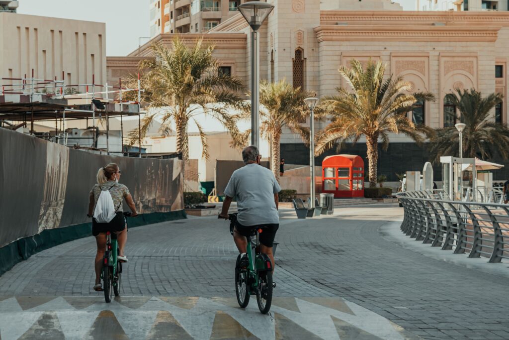 Two people cycling along a waterfront promenade in Dubai Marina under palm trees and modern architecture.
